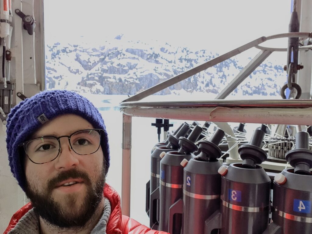 Lowin on the R/V Sikuliaq in Alaska. There are snow-covered mountains behind him. 