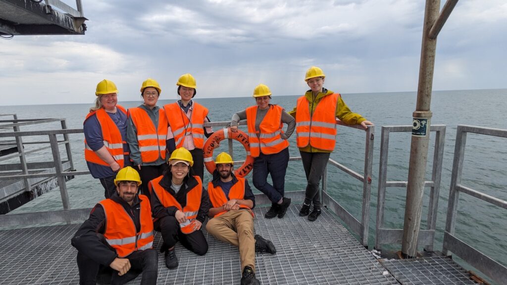 Researchers stand on a metal platform above water. They are all wearing orange protective vests and yellow protective helmets. 